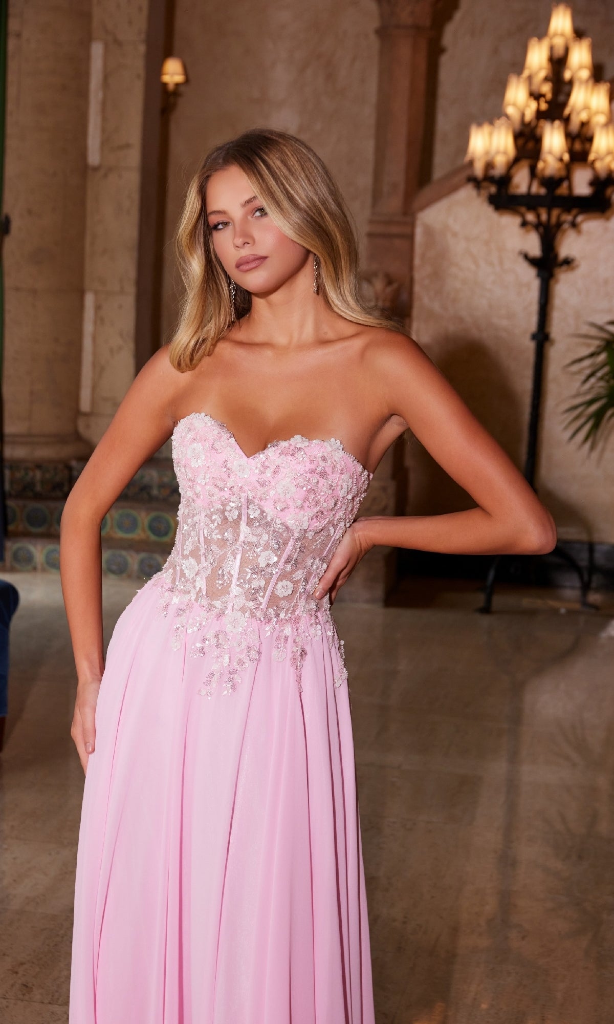 A woman poses indoors wearing the Nox Anabel C1617, a strapless sweetheart prom dress featuring a light pink sheer, floral-embroidered bodice and a flowing skirt.